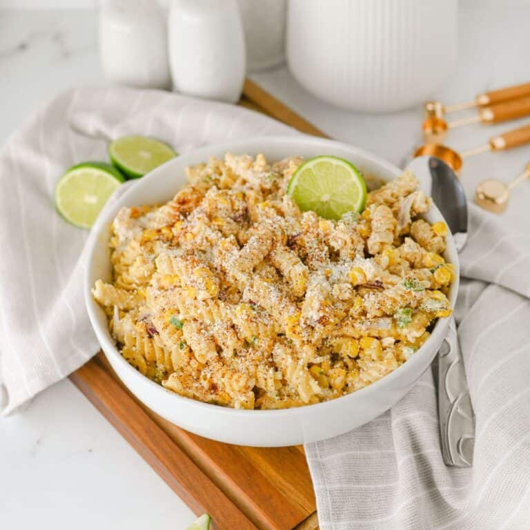 Bowl of pasta mixed with corn, cheese, and seasoning, garnished with lime slices, placed on a light-colored cloth with two spoons, measuring spoons, and salt and pepper shakers in the background.