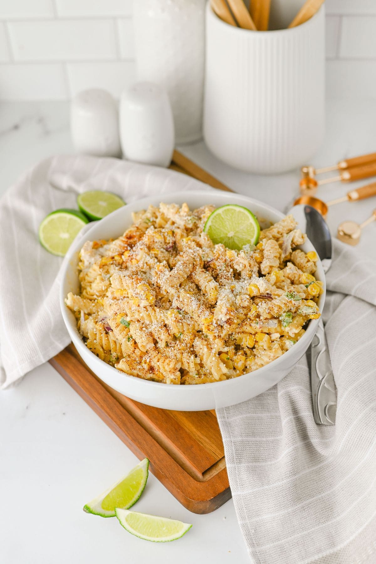 A white bowl filled with pasta mixed with corn and topped with grated cheese and spices, garnished with lime slices. The bowl sits on a wooden board with a gray-striped cloth and utensils in the background.