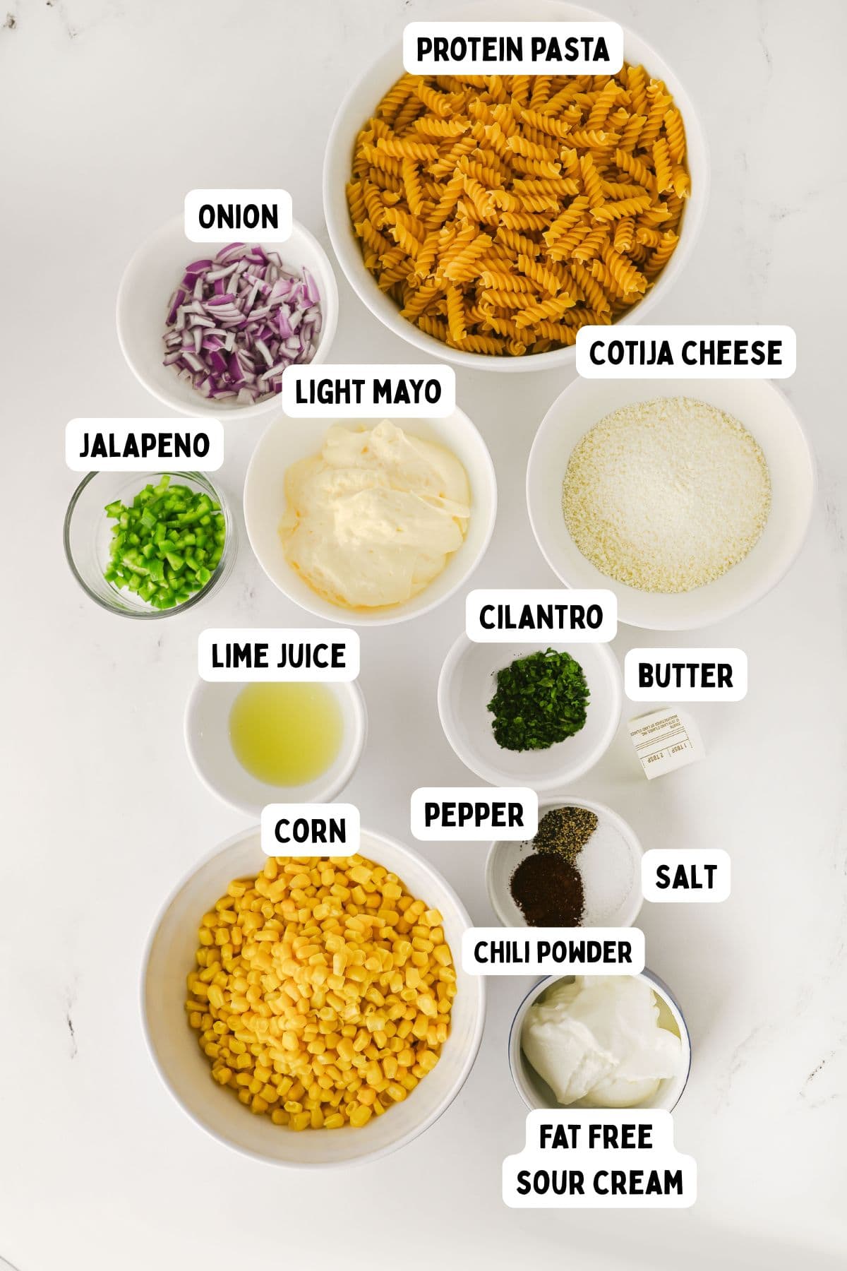 Ingredients for a recipe arranged in bowls on a white surface, labeled as protein pasta, cotija cheese, red onion, jalapeno, light mayo, cilantro, butter, lime juice, pepper, corn, salt, chili powder, and fat-free sour cream.