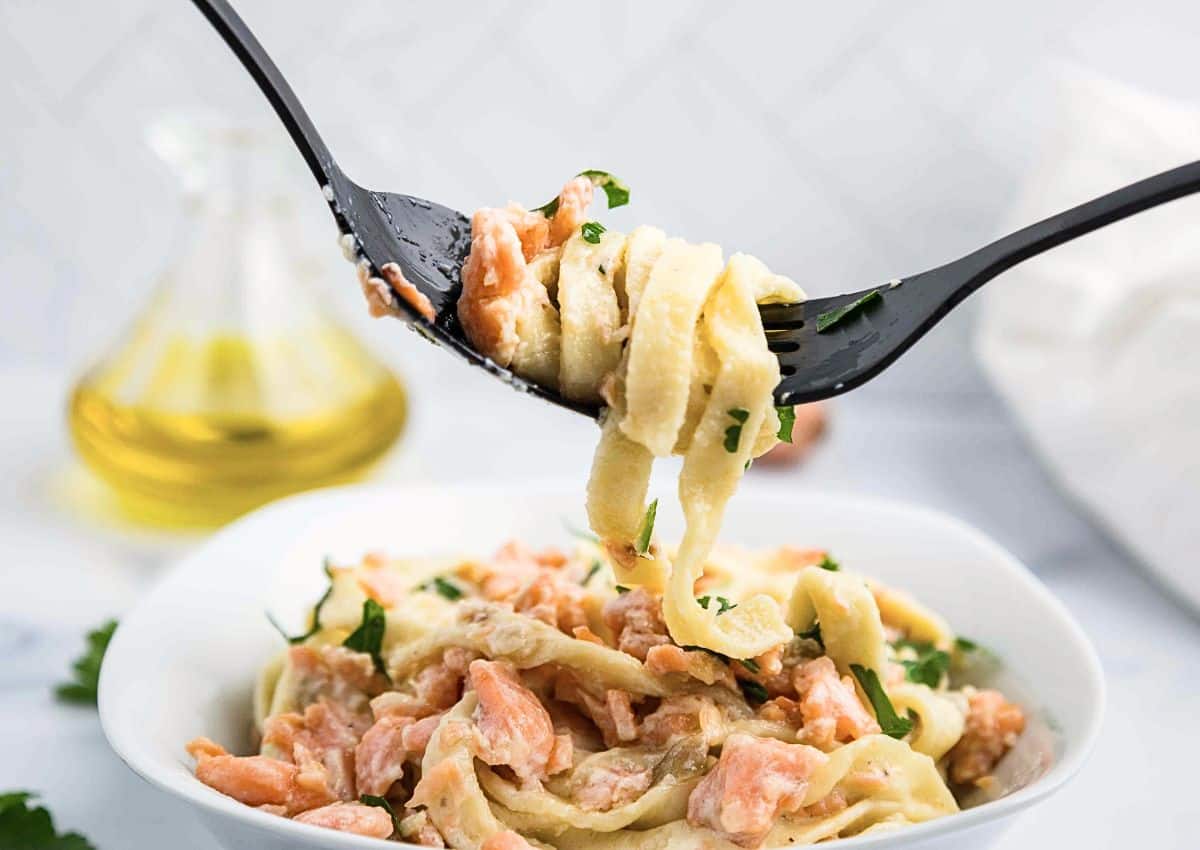 A close-up of a fork lifting creamy fettuccine pasta with pieces of salmon and parsley from a white bowl. A glass bottle of oil is in the background on a white surface.