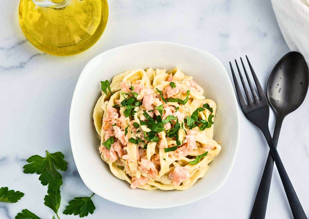 A white bowl of pasta topped with a creamy sauce and chopped herbs sits on a marble surface next to a fork, spoon, a bottle of olive oil, parsley leaves, and a white napkin.