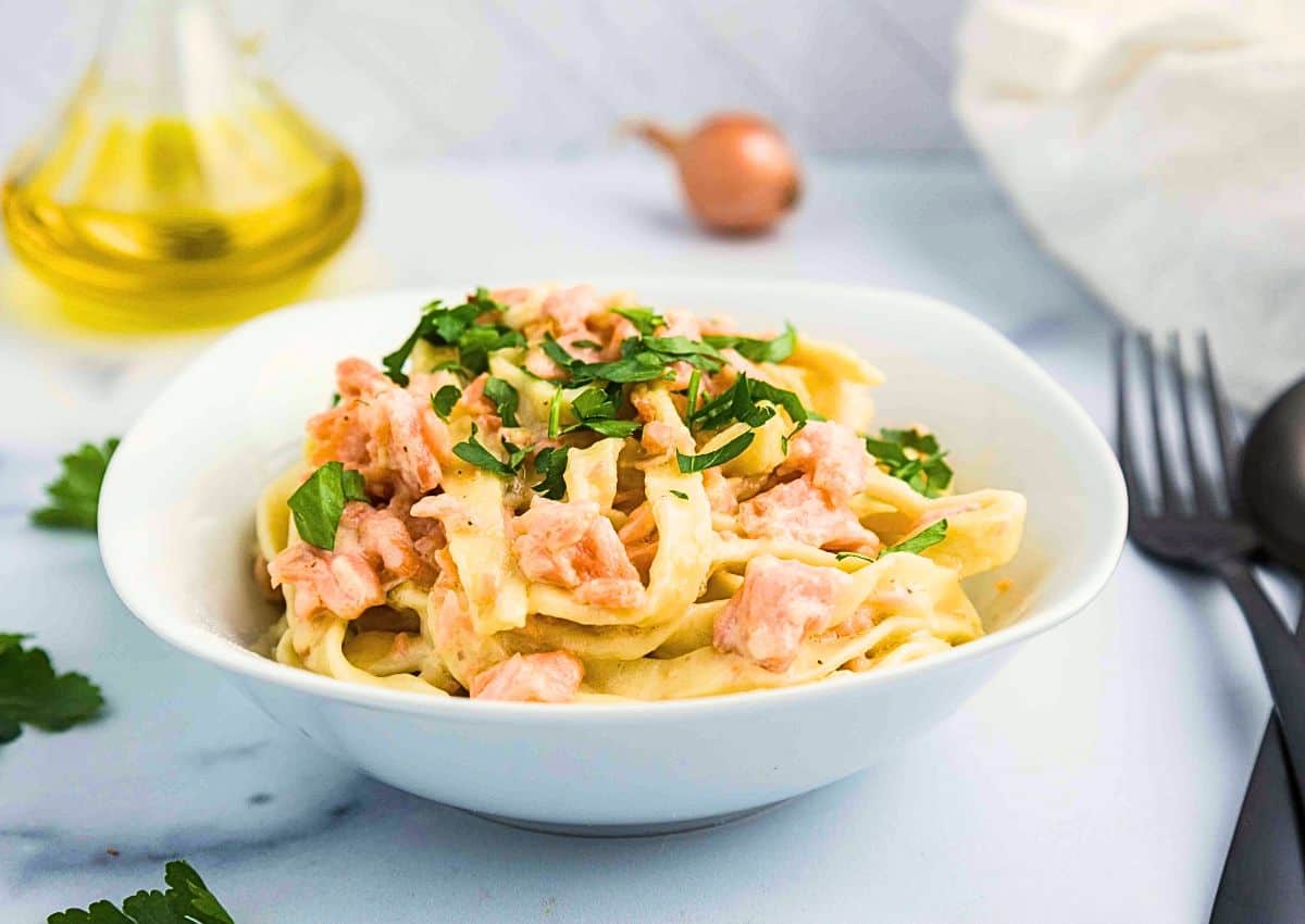 A white bowl filled with fettuccine pasta in a creamy sauce, topped with chunks of salmon and garnished with chopped parsley. A fork, spoon, bottle of oil, and shallot are in the background.