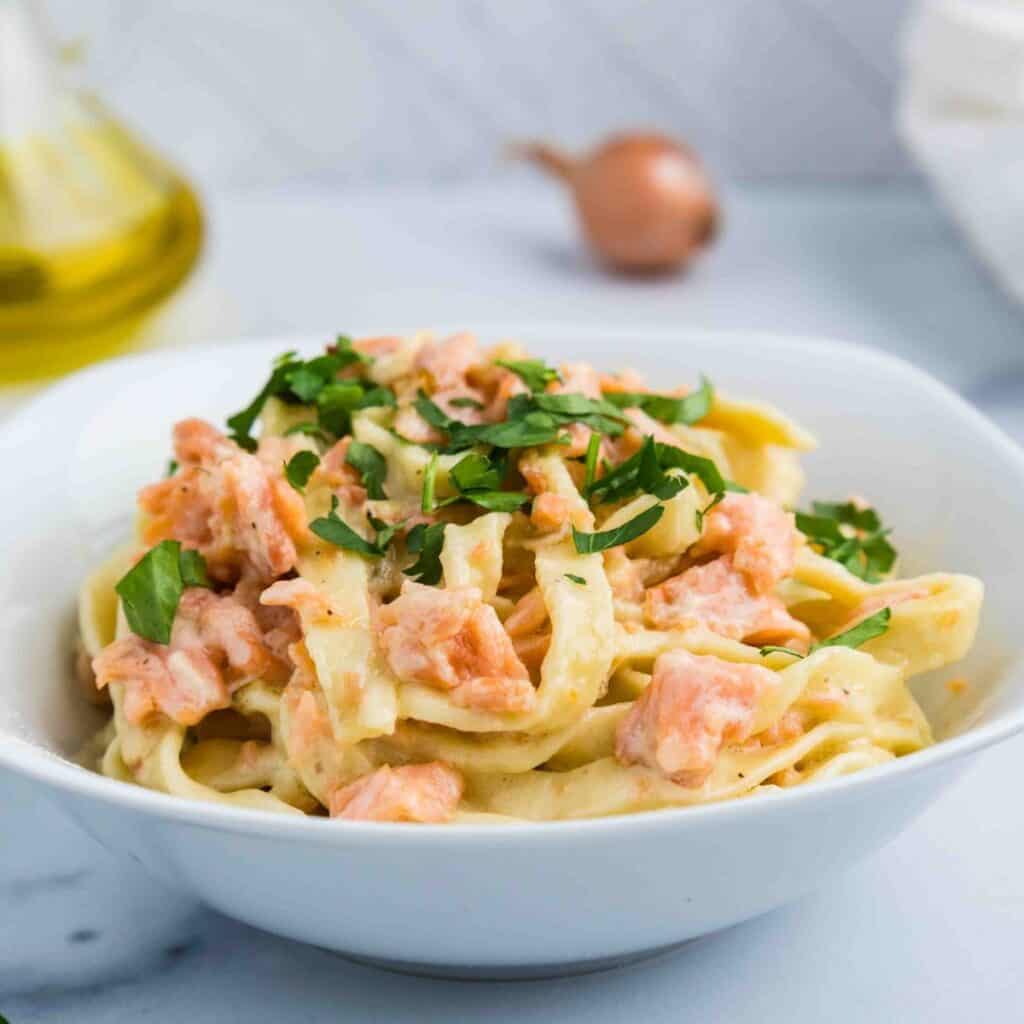 A white bowl filled with Pasta al Salmone—fettuccine pasta topped with creamy sauce, pieces of salmon, and chopped fresh parsley. A bottle of oil and a shallot are blurred in the background.