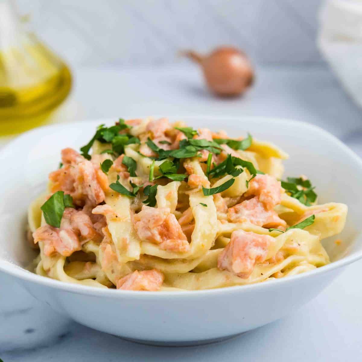 A white bowl filled with Pasta al Salmone—fettuccine pasta topped with creamy sauce, pieces of salmon, and chopped fresh parsley. A bottle of oil and a shallot are blurred in the background.