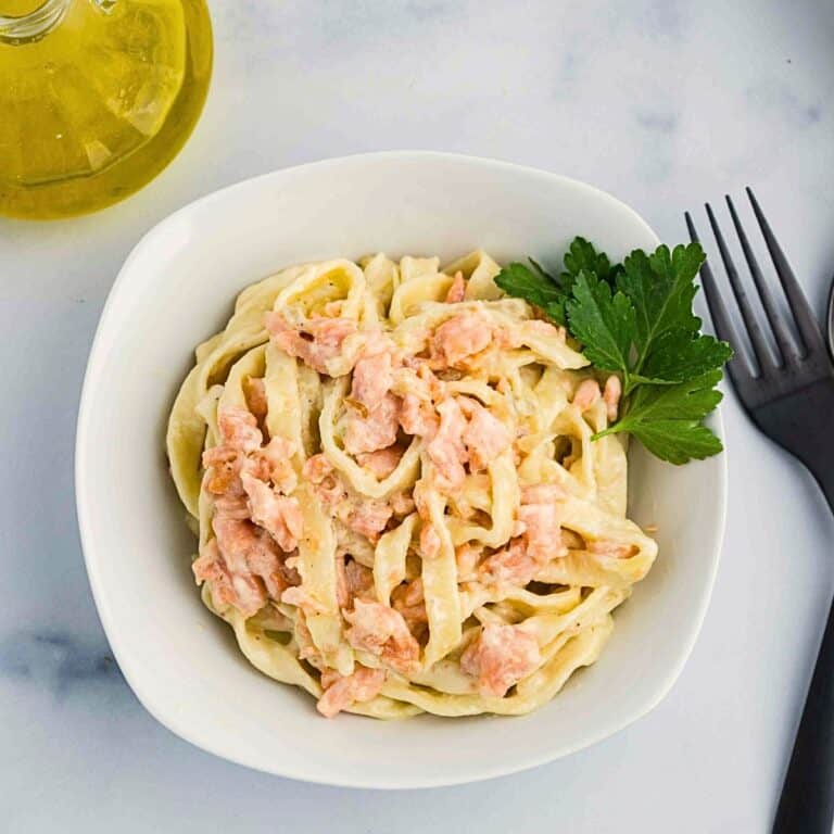A white bowl filled with creamy pasta topped with pieces of salmon and garnished with a sprig of parsley, placed next to a fork, knife, and a bottle of olive oil on a white surface.