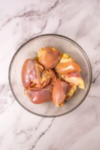 A glass bowl containing six raw, skinless chicken thighs sits on a white marble surface.