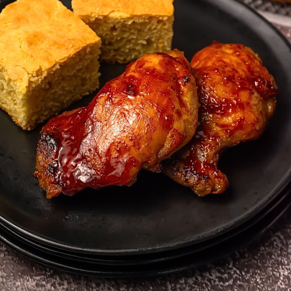 Two pieces of glazed baked chicken and two squares of cornbread are arranged on a black plate. The food is set on stacked plates against a dark background.