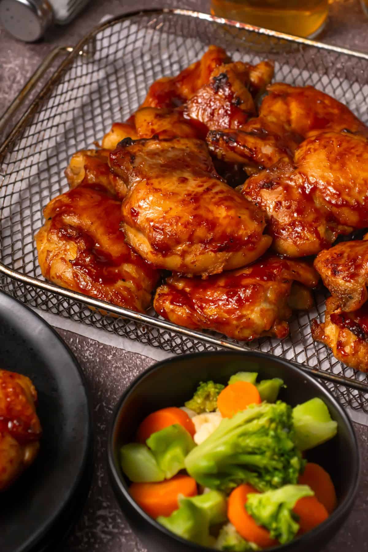 A tray of glazed roasted chicken pieces sits on a wire rack, with a bowl of steamed mixed vegetables including broccoli, carrots, and cauliflower in the foreground. Part of a plate and a drink are partially visible.