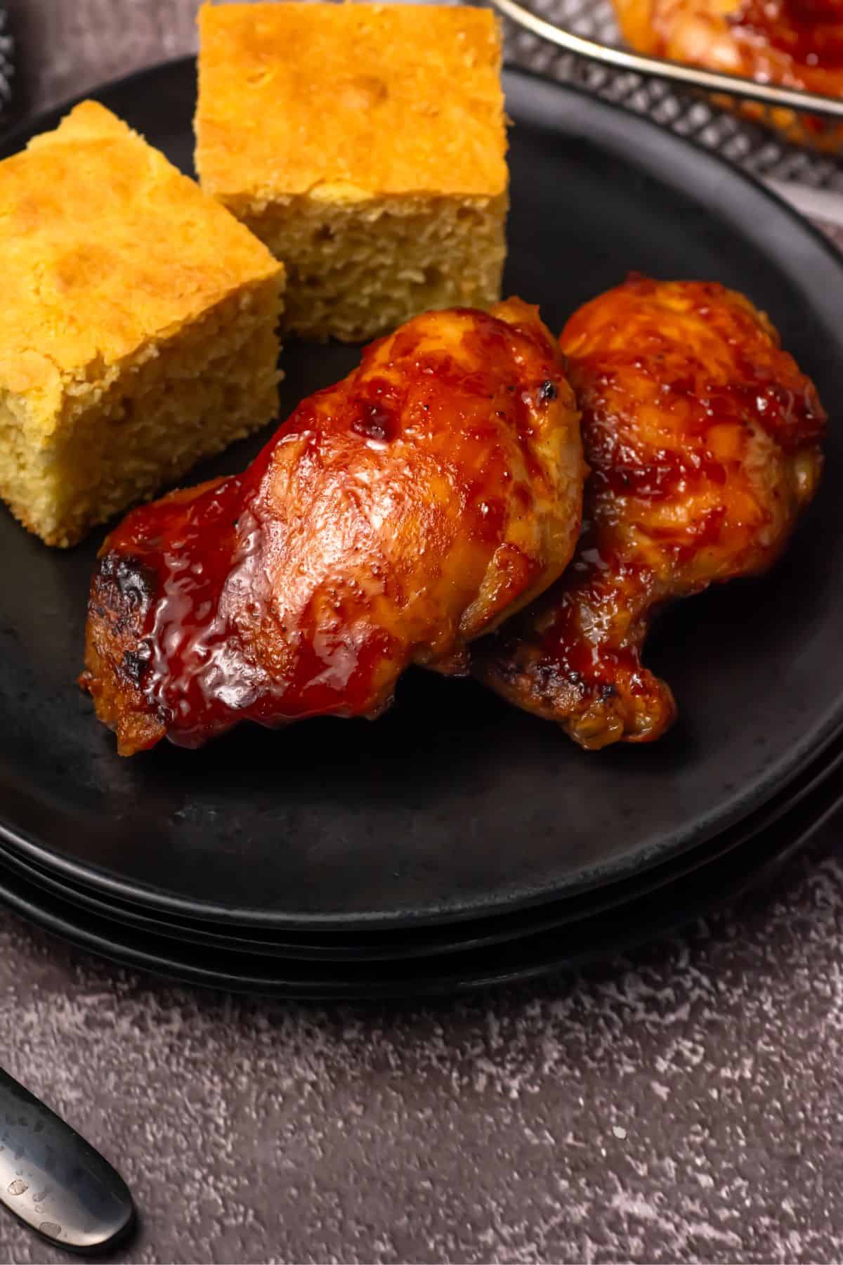 Two pieces of glazed baked chicken and two squares of cornbread are served on a black plate. The food is arranged neatly, and a utensil is visible near the plate on a textured brown surface.