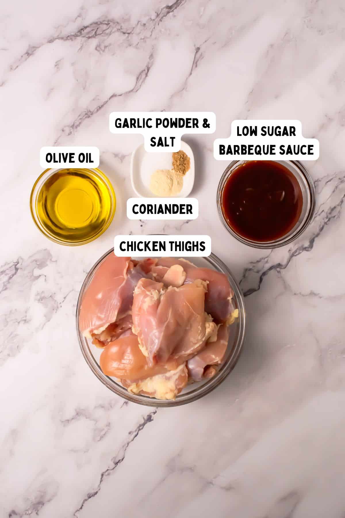 A bowl of raw chicken thighs, a small bowl of olive oil, a small bowl of low sugar barbecue sauce, and small piles of garlic powder, salt, and coriander are arranged on a marble surface. Labels identify each ingredient.