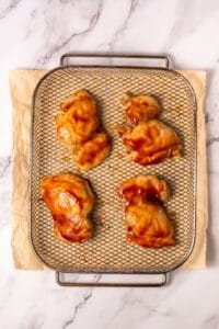 Four pieces of raw, sauce-covered chicken are arranged on a wire rack placed over parchment paper on a marble surface.