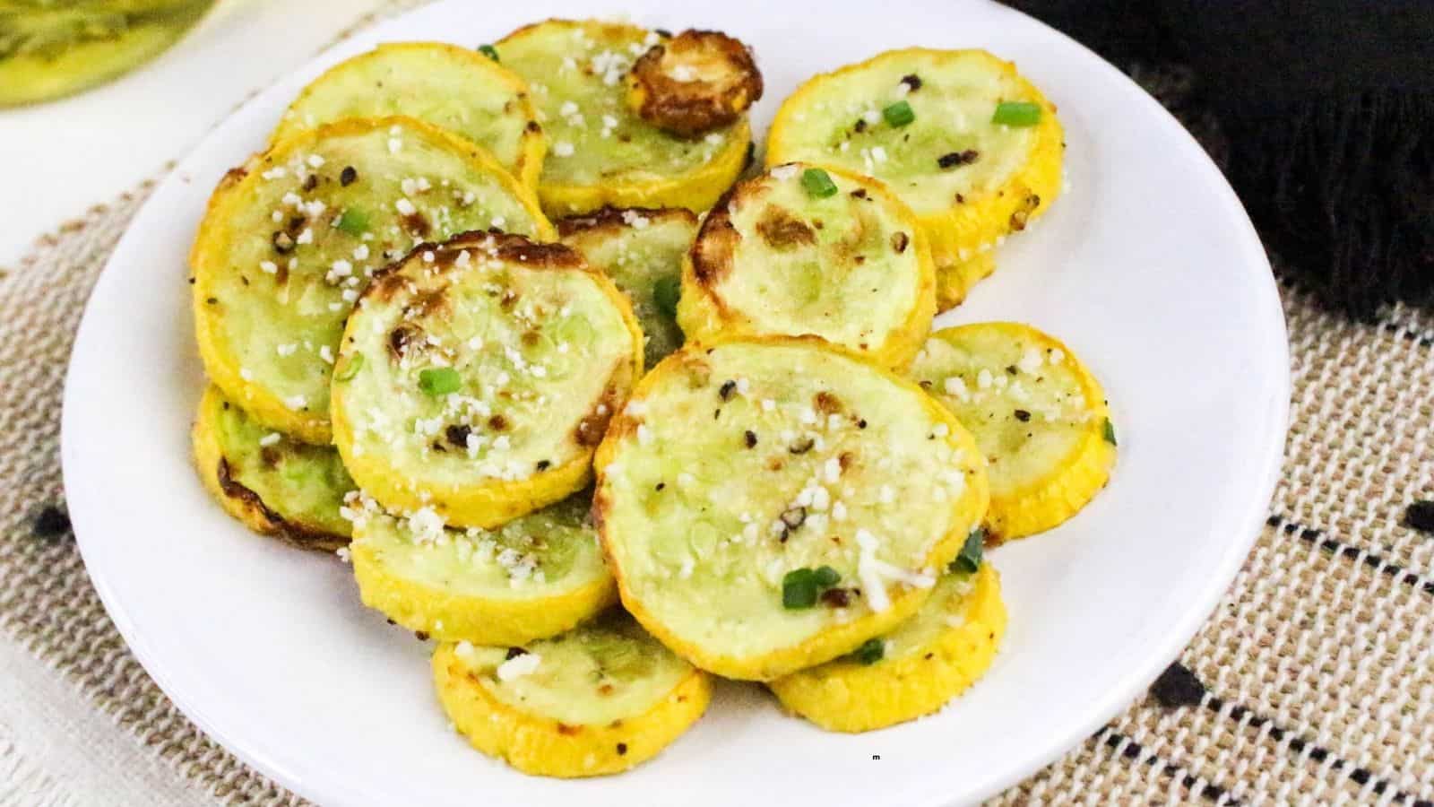 A white plate holds several slices of roasted yellow squash topped with grated cheese and chopped green herbs, resting on a woven beige placemat.