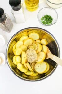 A metal bowl filled with sliced yellow potatoes being mixed with seasonings by a wooden spoon. Surrounding the bowl are containers of salt, pepper, oil, breadcrumbs, chopped herbs, and small bowls of additional spices.