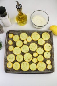 A wire rack holds evenly spaced yellow squash slices, seasoned with herbs. Nearby are a bottle of olive oil, a bowl of breadcrumbs, a salt shaker, a pepper grinder, chopped herbs, and a whole yellow squash.