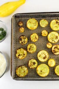 A wire baking rack holds slices of cooked yellow squash with browned spots. Nearby are bowls containing chopped herbs, grated cheese, and a whole yellow squash on a white surface.