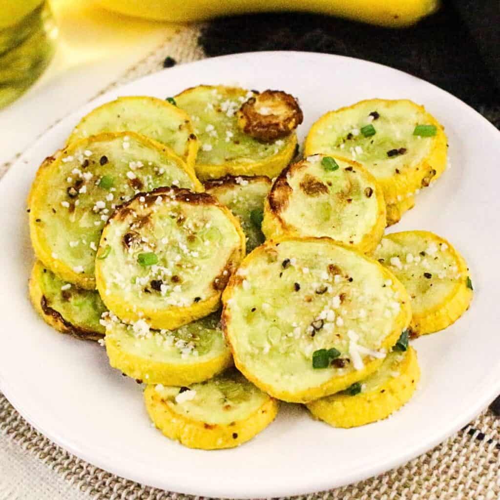 A white plate holds several slices of roasted yellow squash, seasoned with herbs, salt, pepper, and grated cheese. The slices are slightly browned and garnished with chopped green onions.