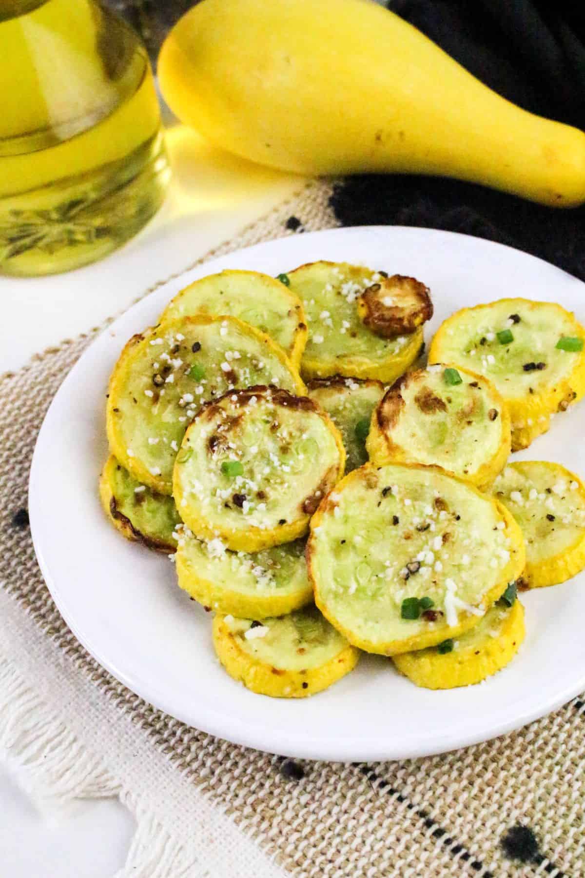 A white plate holds slices of roasted yellow squash topped with seasoning and cheese. A bottle of oil, a whole yellow squash, and a beige textured cloth are visible in the background.