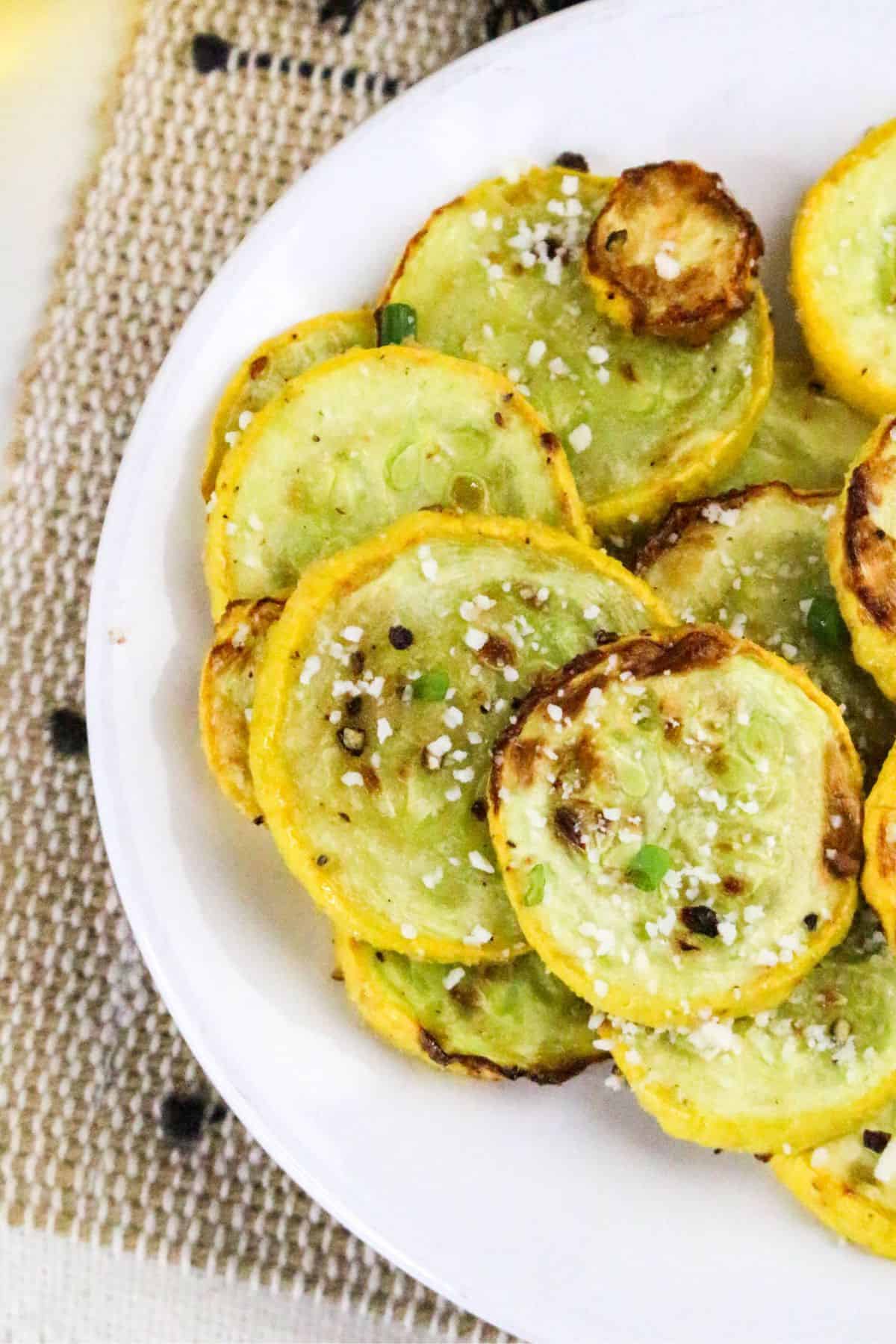 A white plate holds slices of roasted yellow squash, topped with coarse salt, black pepper, and chopped green onions. The plate is on a woven beige placemat with black accents.