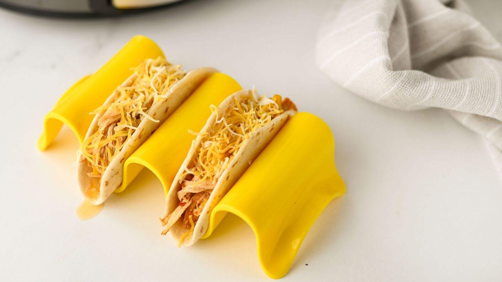Three tacos filled with shredded chicken and topped with shredded cheese are placed in a yellow taco holder on a white surface, with a gray and white striped cloth nearby.