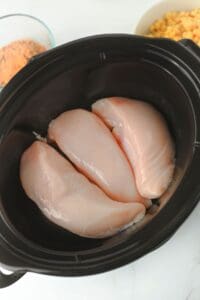 Three raw chicken breasts are placed in a black slow cooker. Bowls containing seasoning and corn are visible in the background on a white surface.
