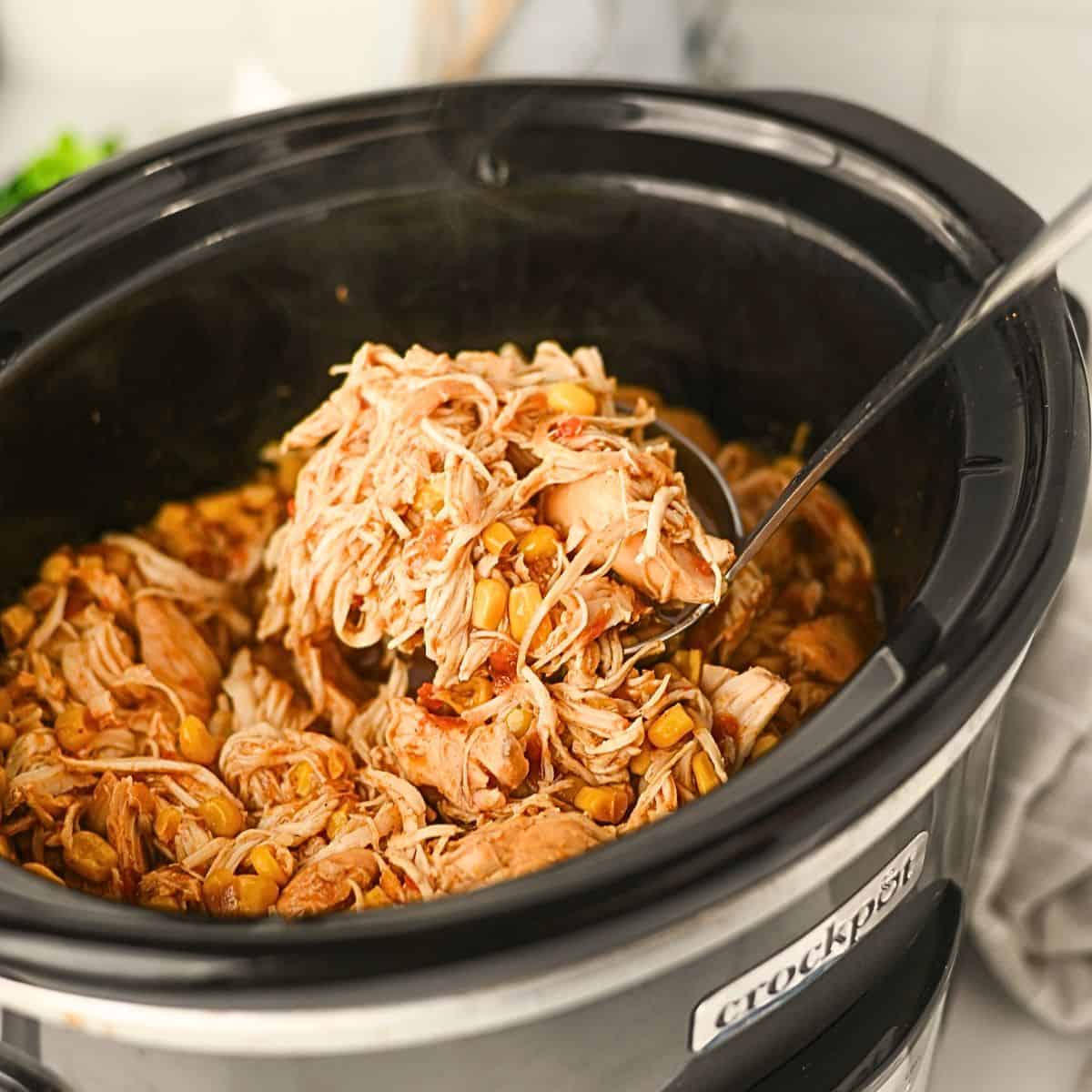 Shredded chicken mixed with beans and spices in a black Crockpot slow cooker, with a metal spoon lifting a portion of the food. Steam is visible rising from the hot dish.