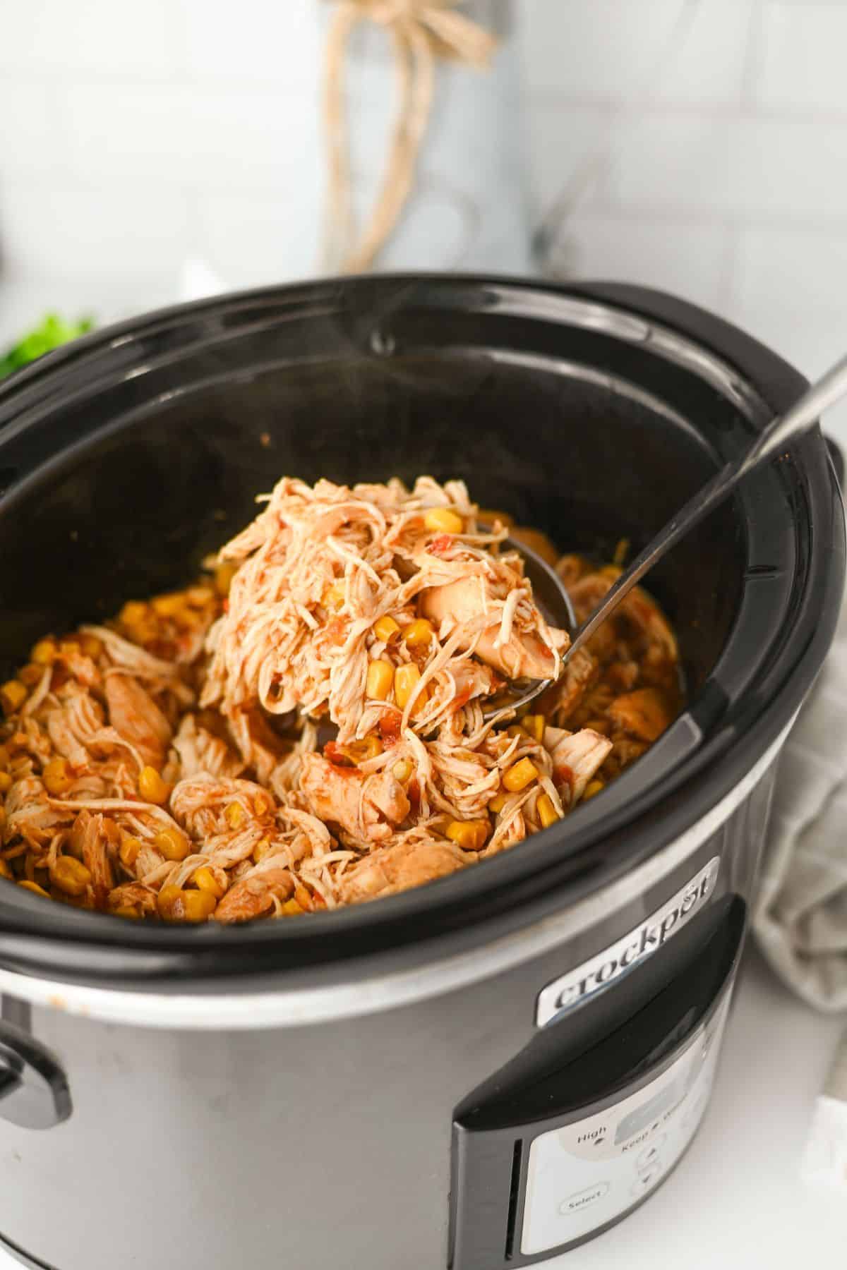 A close-up of shredded chicken with corn and beans inside a black Crockpot slow cooker. A metal serving spoon is lifting a portion of the mixture. The background shows a light kitchen setting.