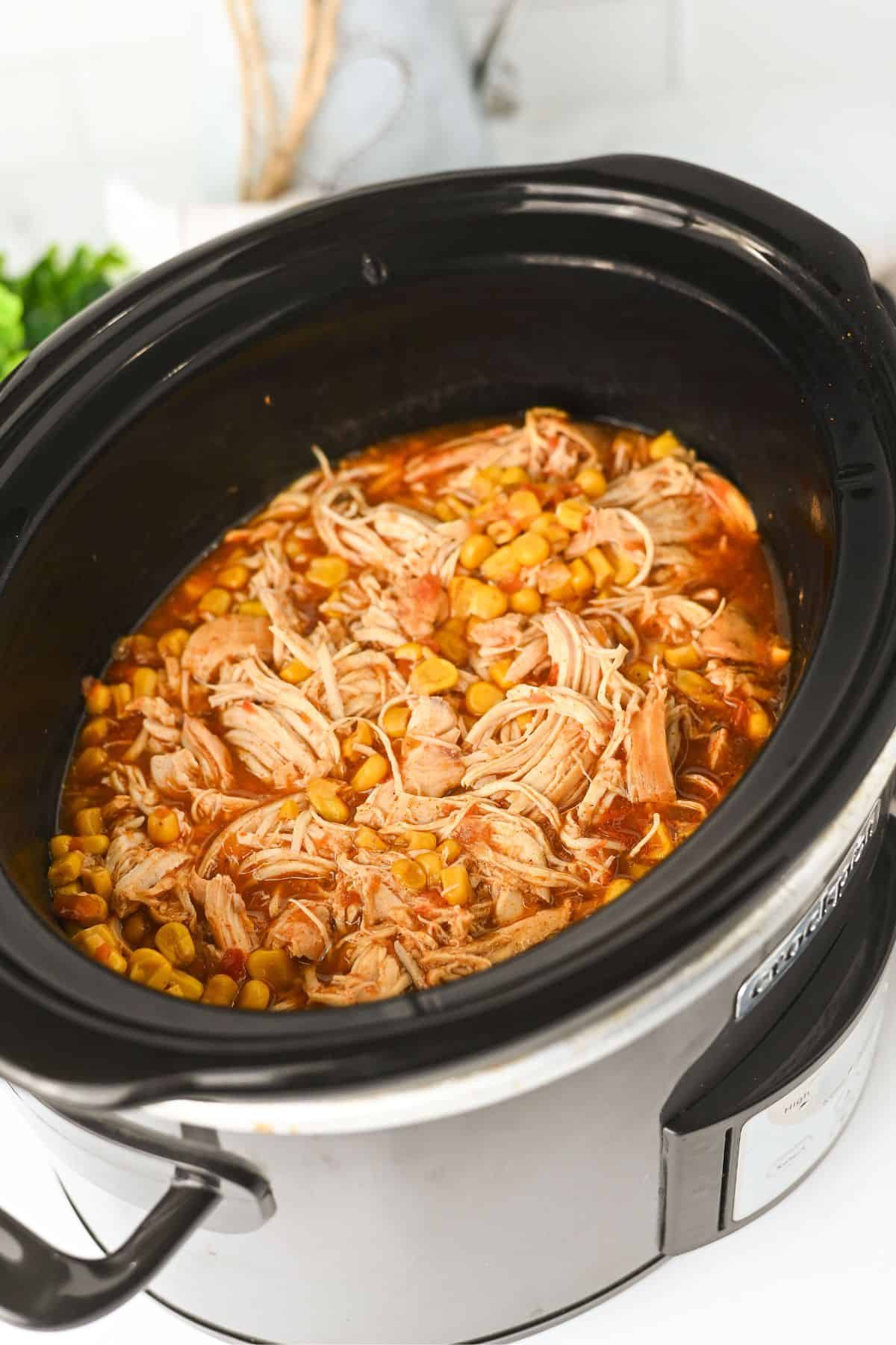 A slow cooker filled with shredded chicken, corn, and a red sauce mixture, sitting on a white countertop with a blurred green garnish and kitchen items in the background.