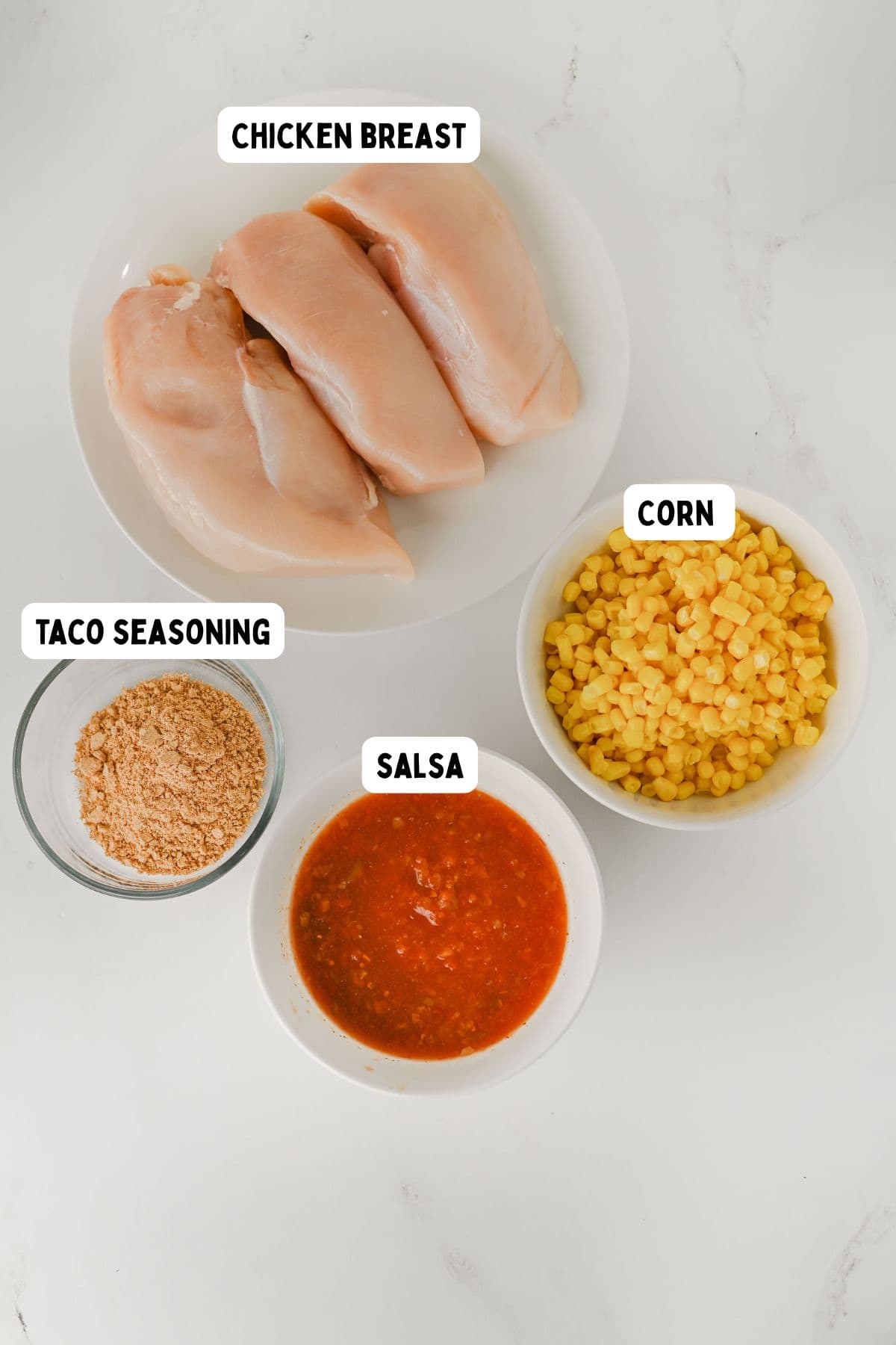 Four bowls on a white surface containing raw chicken breasts, corn, taco seasoning, and salsa. Each bowl is labeled with its contents.