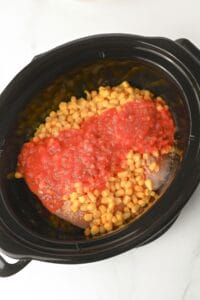 A black slow cooker contains corn kernels and a layer of diced tomatoes or tomato sauce on top of an unseen ingredient, possibly chicken, on a white surface.