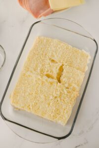 A glass baking dish contains a single layer of sliced bread rolls with the tops removed. Slices of cheese and ham are visible in the background on a white surface.