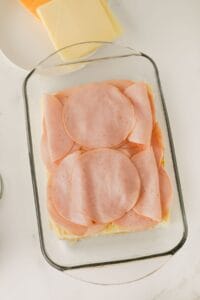 A glass baking dish filled with layered slices of ham on top of bread. In the background, a plate with slices of white and yellow cheese is partially visible. The setting is on a white countertop.