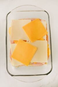 A glass baking dish contains layered slices of white and yellow cheese on top of ham and bread, prepared for baking.