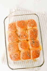 A glass baking dish filled with twelve golden-brown slider buns topped with seasoning, resting on a folded white and gray striped cloth on a white surface.