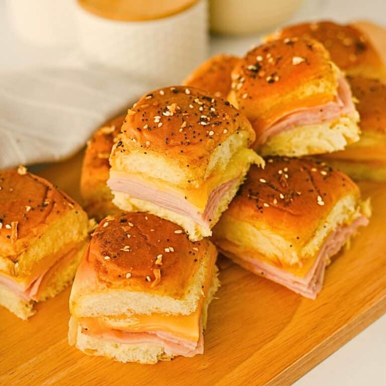 A group of ham and cheese sliders with seasonings on top of golden buns, arranged on a wooden serving tray. A white napkin and ceramic dishes are visible in the background.