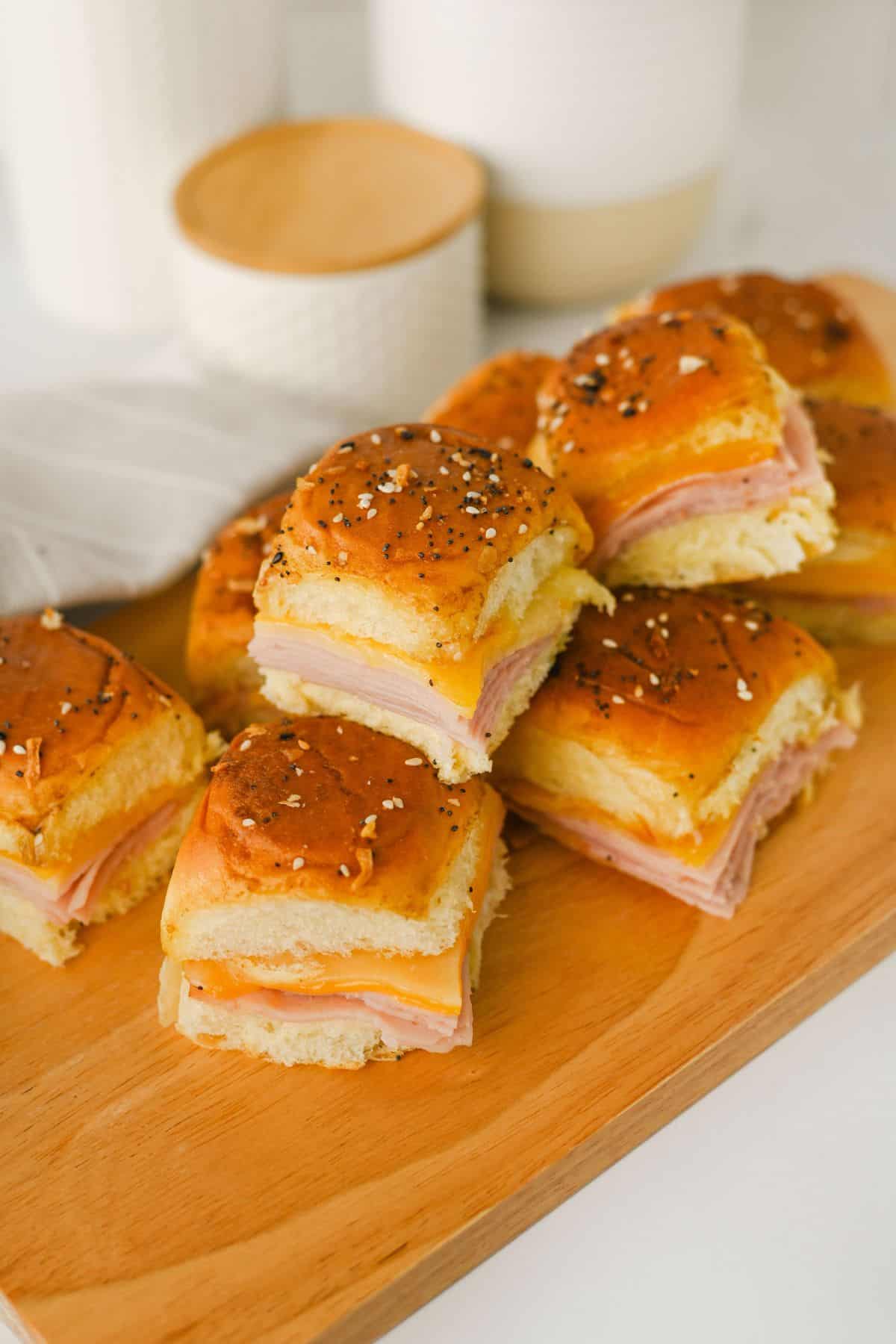 A group of small ham and cheese sliders with seasoned tops are arranged on a wooden cutting board. The background shows a folded napkin and white containers.
