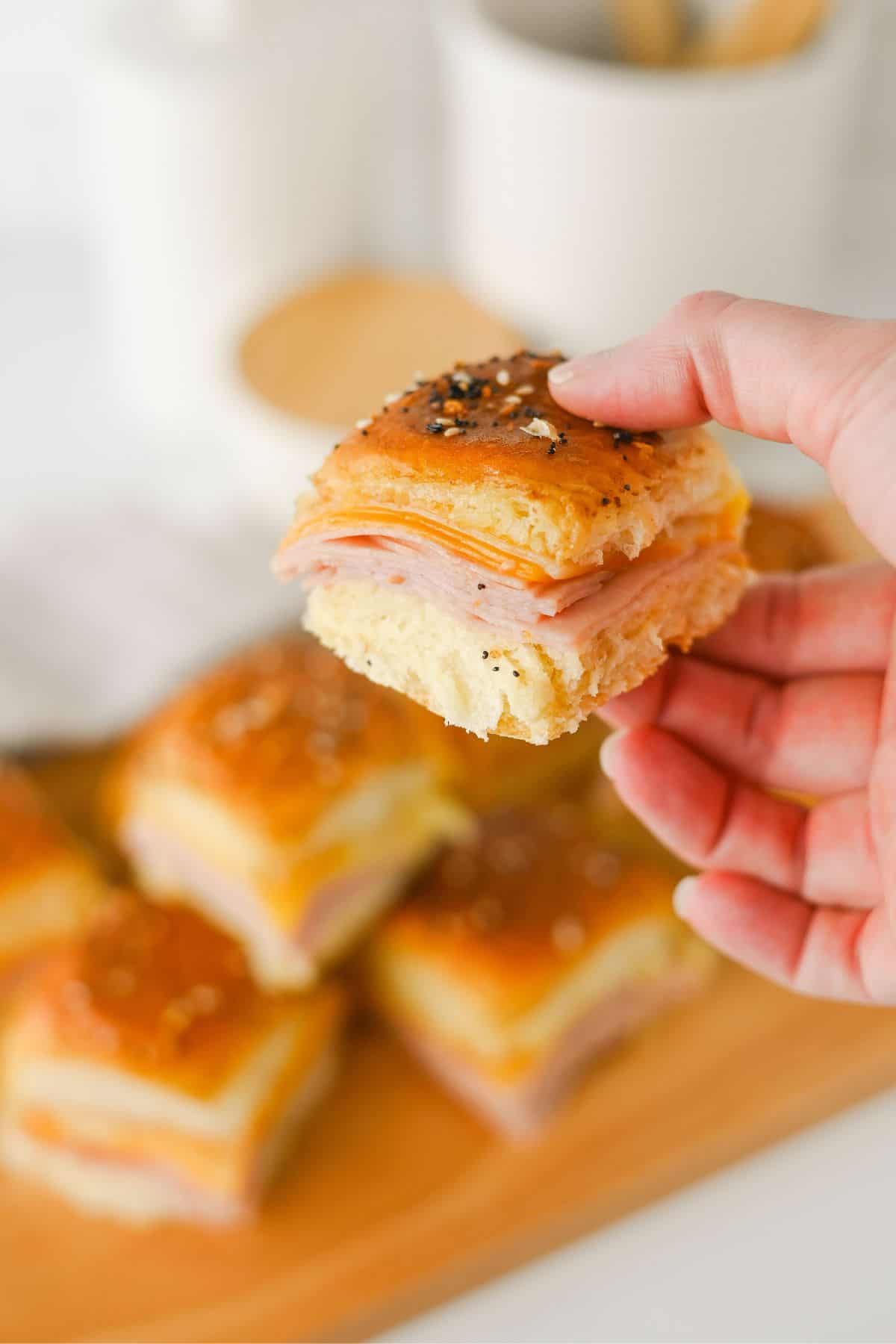 A hand holds a small sandwich made of layered ham and cheese on a soft, golden bun topped with seasoning. More sandwiches are arranged on a wooden board in the background.