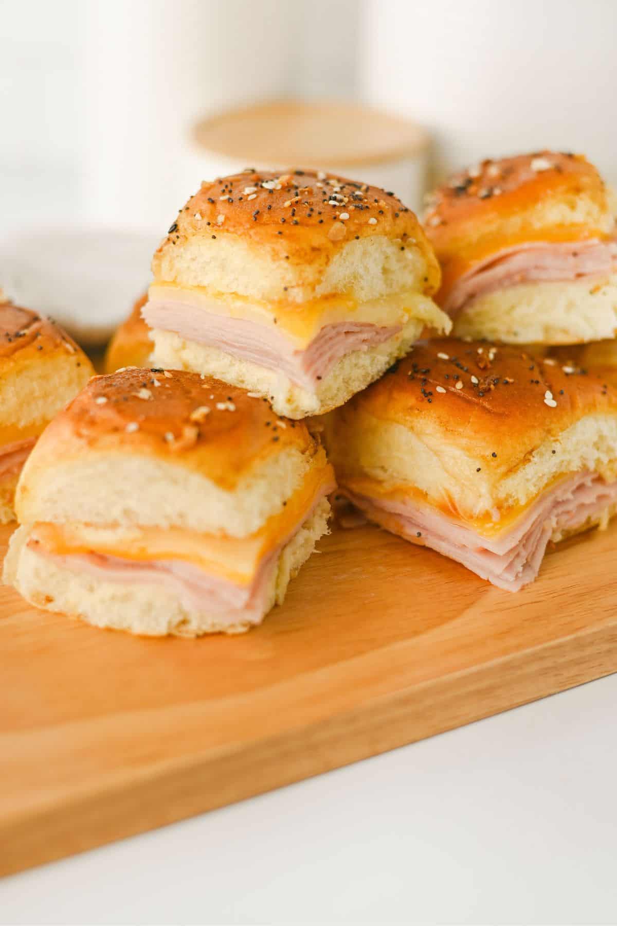A wooden board topped with several small sandwiches made with soft rolls, deli meat, and cheese, garnished with seasoning on the buns.