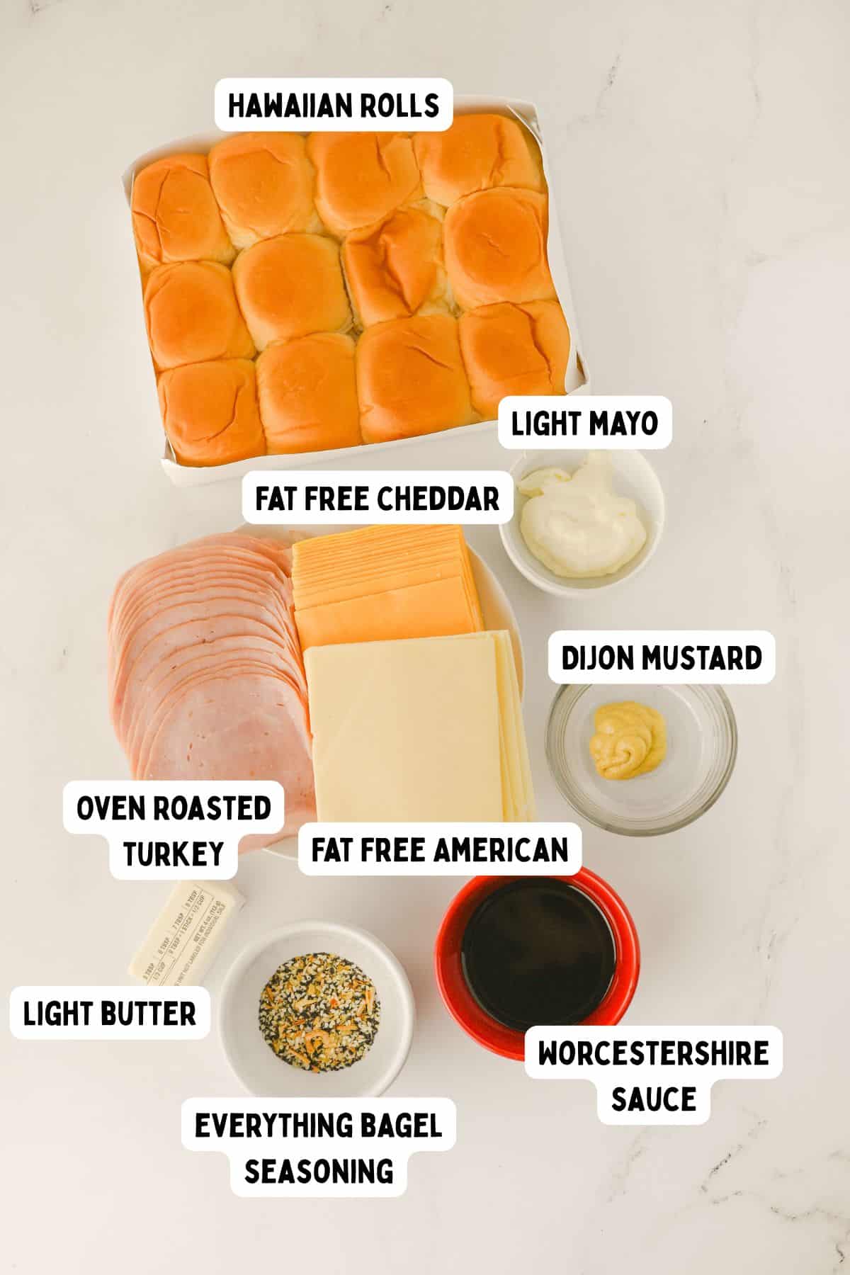 A top-down view of ingredients on a white surface: Hawaiian rolls, fat free cheddar, fat free American cheese, oven roasted turkey slices, light mayo, Dijon mustard, Worcestershire sauce, light butter, and everything bagel seasoning.