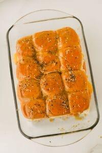 A glass baking dish filled with twelve golden brown slider buns topped with a glossy glaze and sprinkled with poppy seeds and seasoning, resting on a white surface.