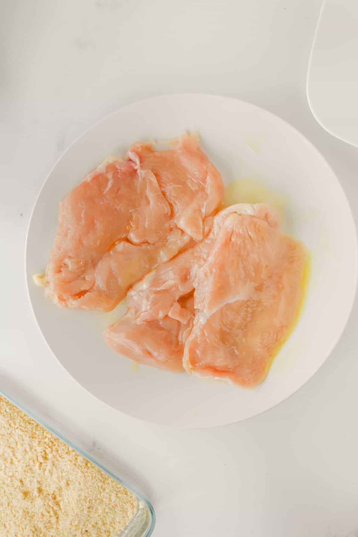 A white plate with three raw, boneless chicken breast fillets drizzled with oil sits on a white surface. A glass container with breadcrumbs is partially visible in the lower left corner.
