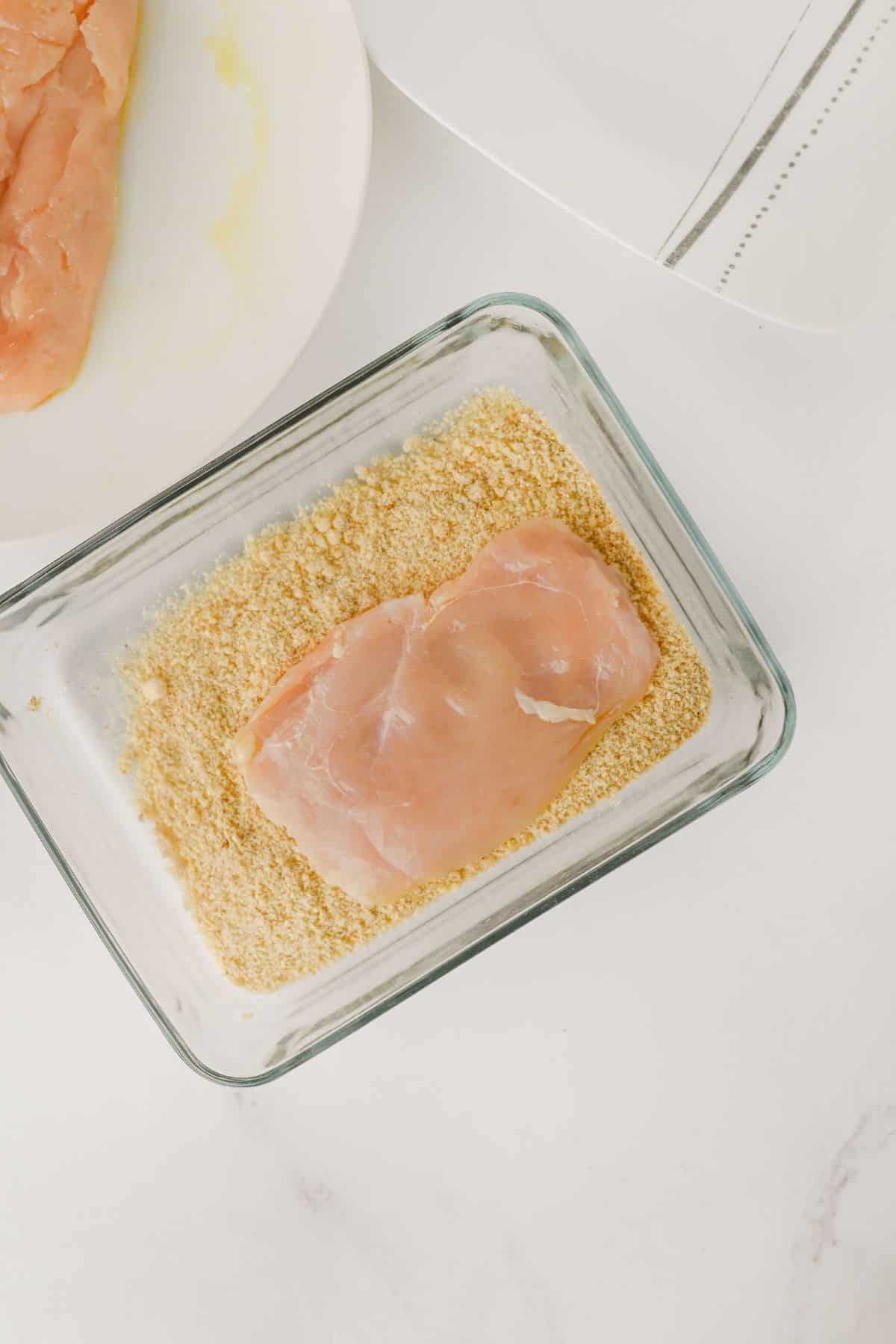 A raw chicken breast is placed in a glass container filled with breadcrumbs. A plate with another piece of raw chicken is partially visible in the background. The scene is set on a white surface.