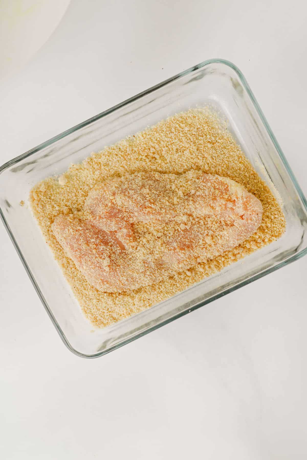 A raw chicken breast coated in breadcrumbs sits in a glass dish on a white surface.