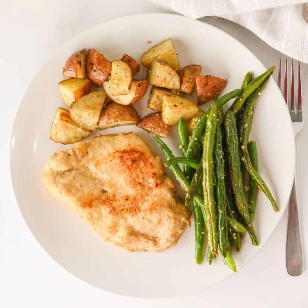 A white plate with baked breaded chicken breast, roasted potato pieces, and cooked green beans. A fork is partially visible to the right of the plate, with a white cloth at the top edge.
