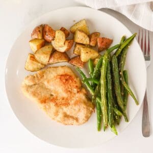 A white plate with baked breaded chicken breast, roasted potato pieces, and cooked green beans. A fork is partially visible to the right of the plate, with a white cloth at the top edge.