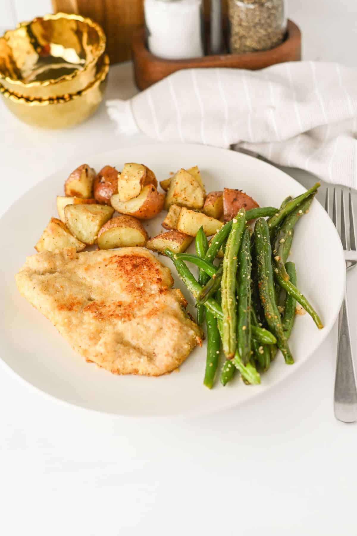 A white plate with breaded chicken cutlet, roasted potato pieces, and green beans. A fork is beside the plate, with bowls and seasonings in the background.