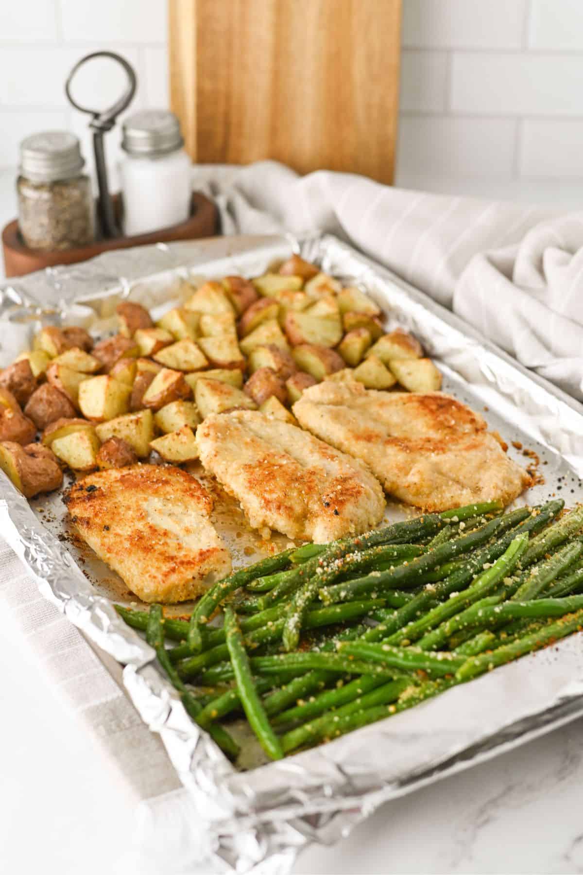 A foil-lined baking sheet with three breaded chicken breasts, roasted potato cubes, and green beans, all seasoned and baked. Salt and pepper shakers and a cutting board are in the background.