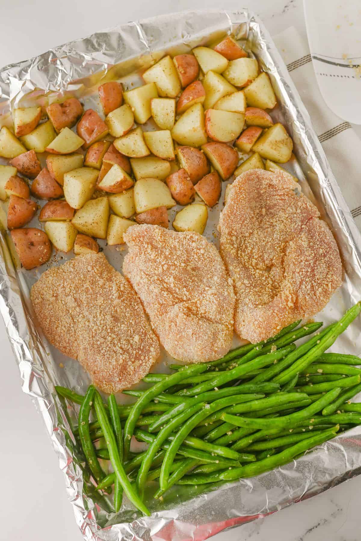 A foil-lined baking sheet with three breaded chicken breasts, chopped seasoned red potatoes, and fresh green beans arranged in separate sections, ready to be baked.