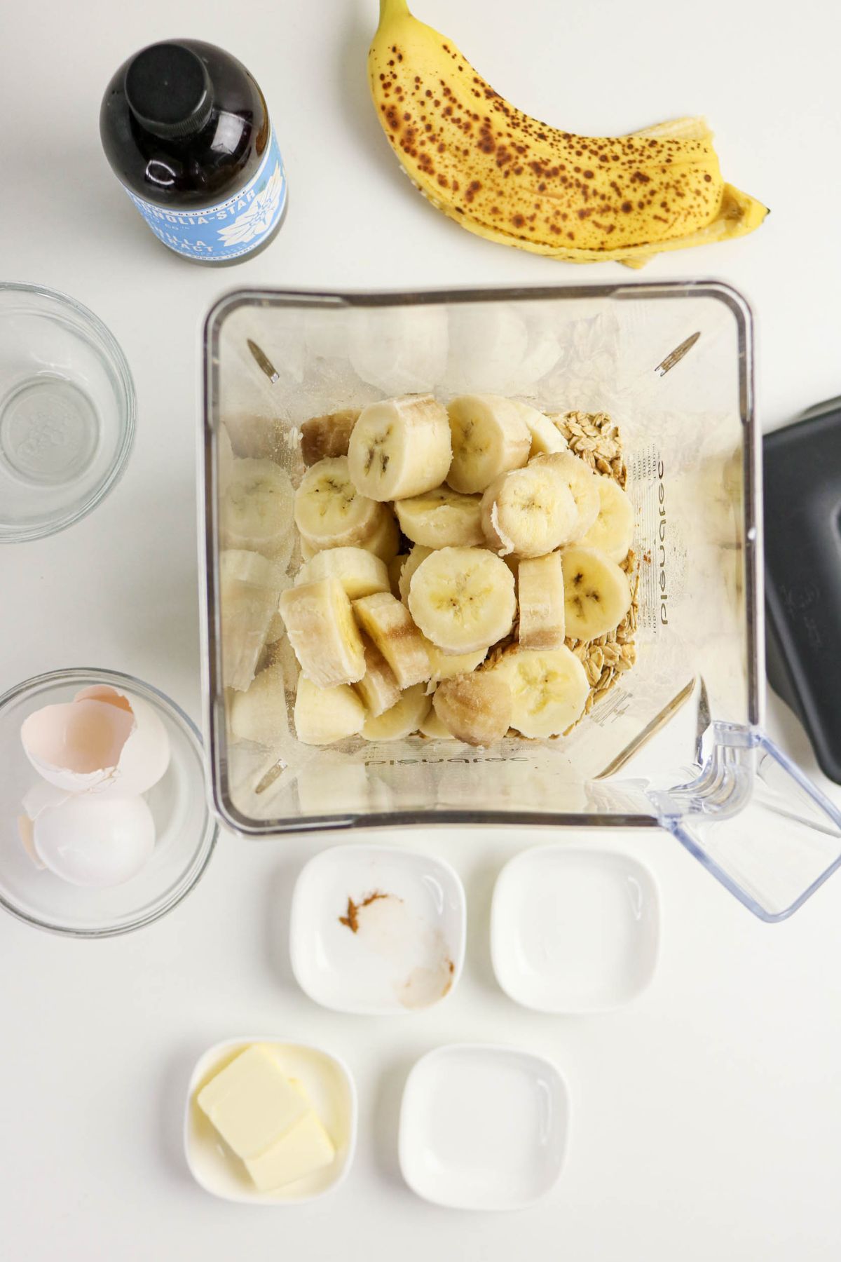 A blender filled with sliced bananas and oats is surrounded by a ripe banana, vanilla extract, empty glass bowls, eggshells, a pat of butter, a small amount of vanilla, and other small white bowls on a white surface.