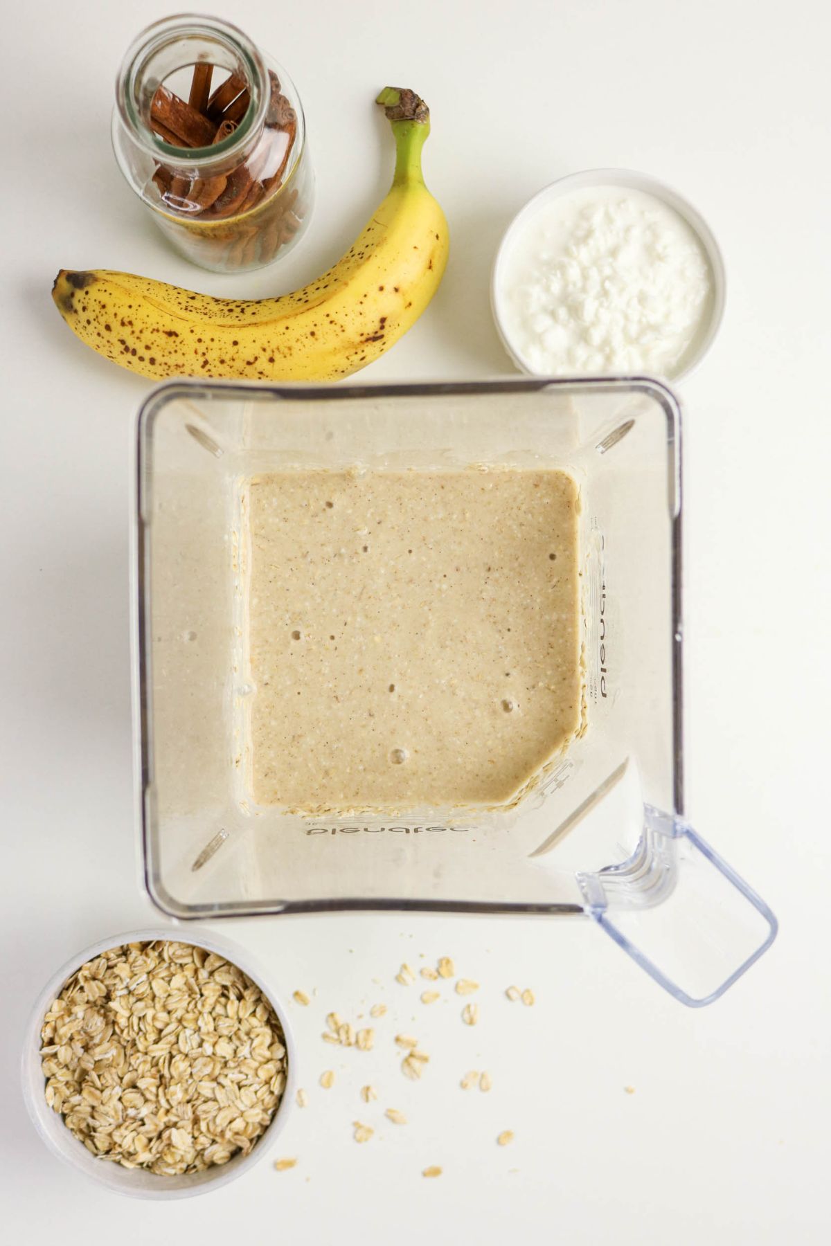 A blender filled with a beige smoothie mixture is on a white surface, surrounded by a banana, a jar of almonds, a bowl of rolled oats, and a bowl of white yogurt. Some oats are scattered nearby.