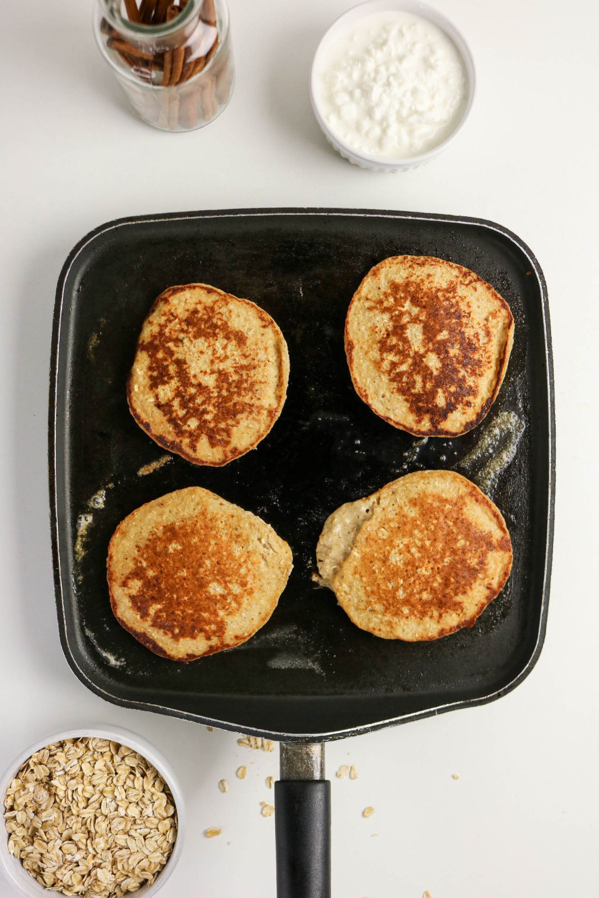 Four golden-brown pancakes are cooking on a square black griddle. Nearby are bowls containing oats and cottage cheese, along with a jar of cinnamon sticks. The items are placed on a white surface.